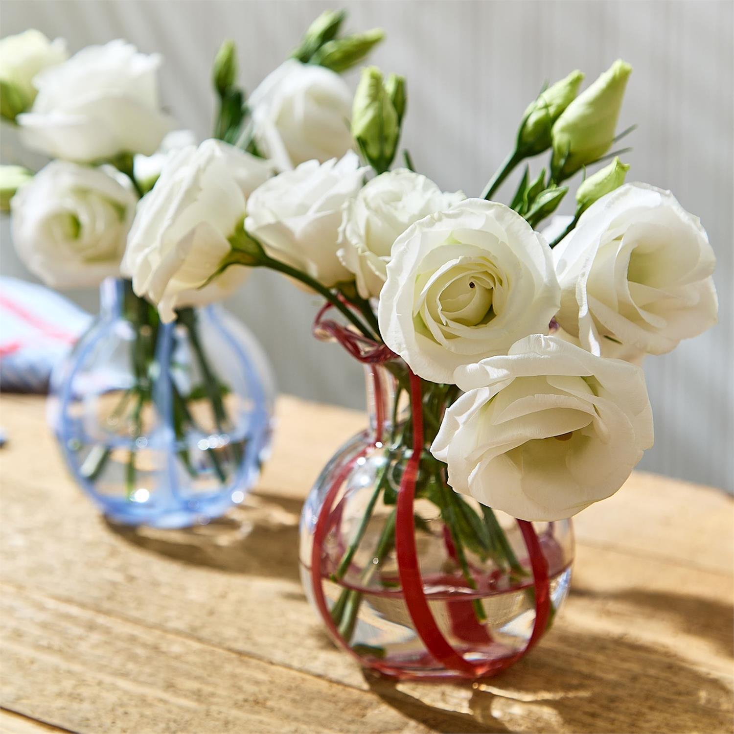Two glass vases holding white Lisianthus flowers and green buds on a sunlit wooden table, one vase intricately striped with red and the other with blue