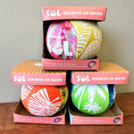 SOL Bounces on Water product photograph displaying three stacked premium foam balls in their boxes on a wooden surface, highlighting their change-color-in-the-sun feature and vibrant patterns including pink Waboba, yellow pineapple, orange palm fronds, and green palm trees