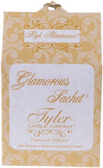 Tyler Candle Company's "High Maintenance" Glamorous Sachet product box features a rich golden yellow background with elegant white scrollwork patterns, a prominent white label with "Glamorous Sachet" in ornate gold script, and a decorative braided gold hanging loop at the top