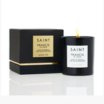 Saint Francis of Assisi brand luxury candle with a lit flame in a black vessel and its matching black box featuring gold accents and "Heavenly Tree" scent details, displayed on a reflective white surface