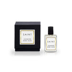 Product photograph featuring a black SAINT Jasmine and Oud perfume box with a gold-bordered label and a matching clear glass bottle with a black cap, both standing on a reflective white surface