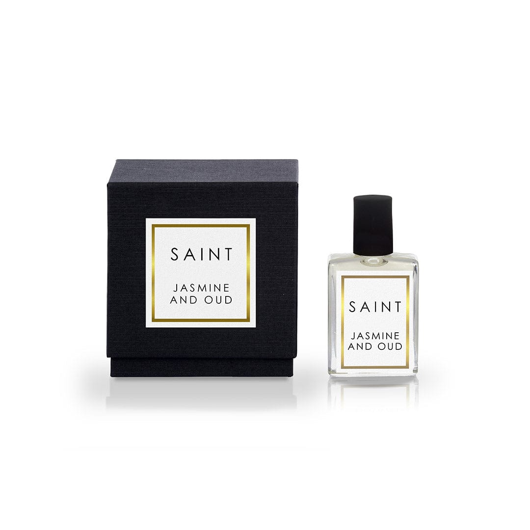Product photograph featuring a black SAINT Jasmine and Oud perfume box with a gold-bordered label and a matching clear glass bottle with a black cap, both standing on a reflective white surface