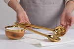 A person in a grey apron presents two glossy golden serving utensils, one slotted, above a wood-grain textured bowl with a golden rim resting on a white napkin