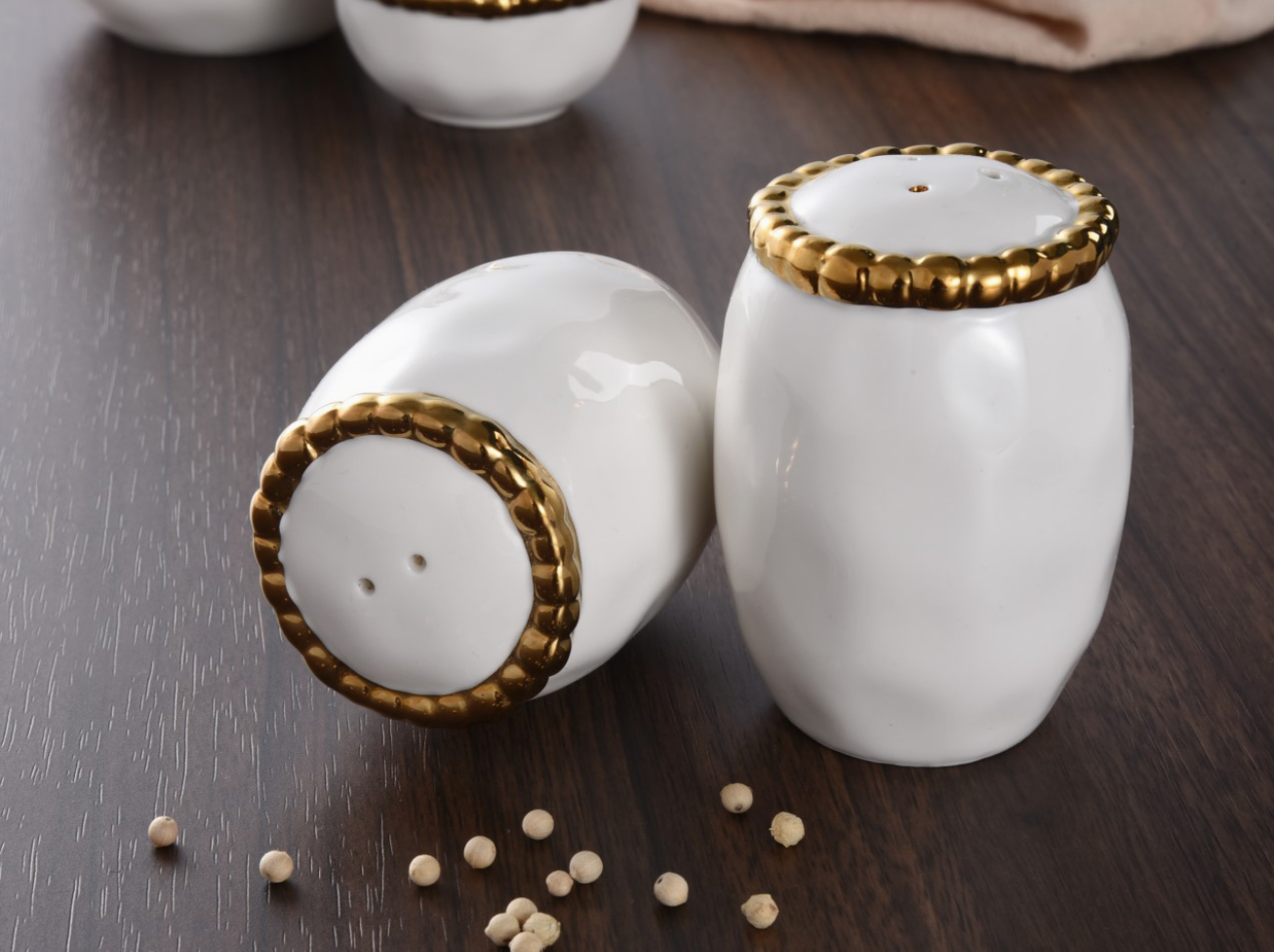 Two white ceramic shakers with decorative gold braided trim, one upright and one lying on a dark wooden tabletop with scattered white peppercorns