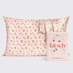 Kitsch satin pillowcase with a vibrant red cherry pattern on a pale pink background next to its coordinating pink product box displaying the brand logo