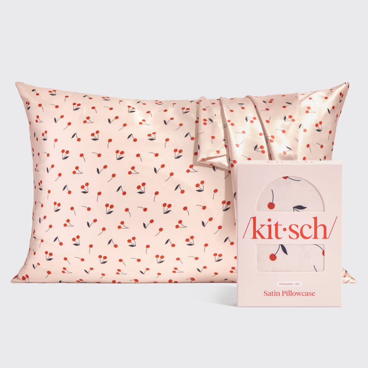 Kitsch satin pillowcase with a vibrant red cherry pattern on a pale pink background next to its coordinating pink product box displaying the brand logo
