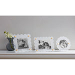 Three scalloped clear acrylic photo frames with gold posts displaying black and white family images arranged on a white shelf next to a grey vase with white blossoms