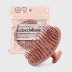 Kitsch product image of a blush pink silicone scalp exfoliator brush with prominent bristles and a handle, positioned in front of its clear packaging displaying 'recycled plastic' and 'woman-owned business' labels and 'scalp exfoliator' text