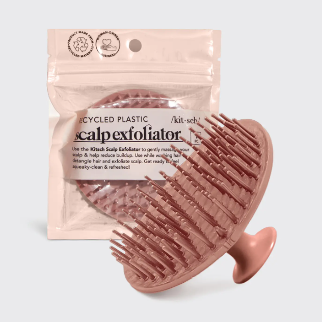 Kitsch product image of a blush pink silicone scalp exfoliator brush with prominent bristles and a handle, positioned in front of its clear packaging displaying 'recycled plastic' and 'woman-owned business' labels and 'scalp exfoliator' text