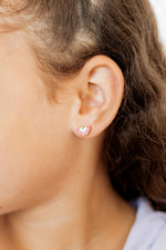 Close-up photograph of a child's ear adorned with a small gold butterfly stud earring featuring intricate pink and white enamel detailing amidst soft brown wavy hair