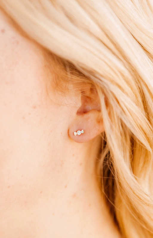 Photograph of a woman's ear with wavy blonde hair wearing a small gold-toned candy wrapper shaped stud earring featuring a central round diamond