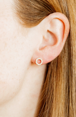 Close-up of a person's earlobe adorned with a delicate gold-toned open circle stud earring featuring tiny sparkling diamonds set against fair skin and flowing light brown hair