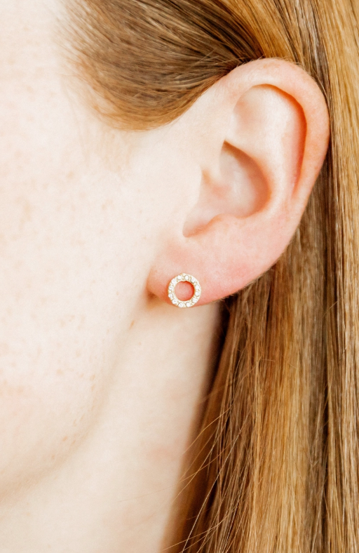 Close-up of a person's earlobe adorned with a delicate gold-toned open circle stud earring featuring tiny sparkling diamonds set against fair skin and flowing light brown hair