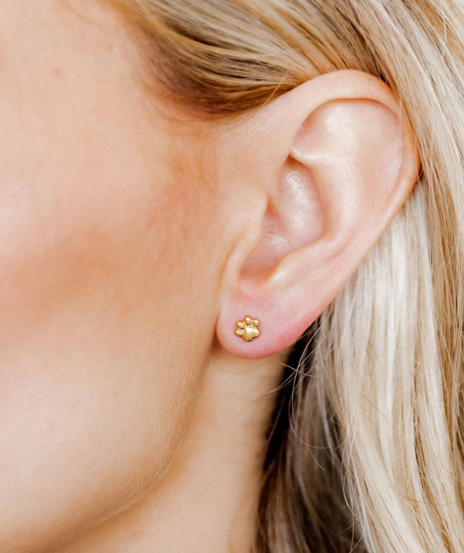 Photograph of a woman's ear adorned with a small golden paw print stud earring against fair skin with blonde hair visible