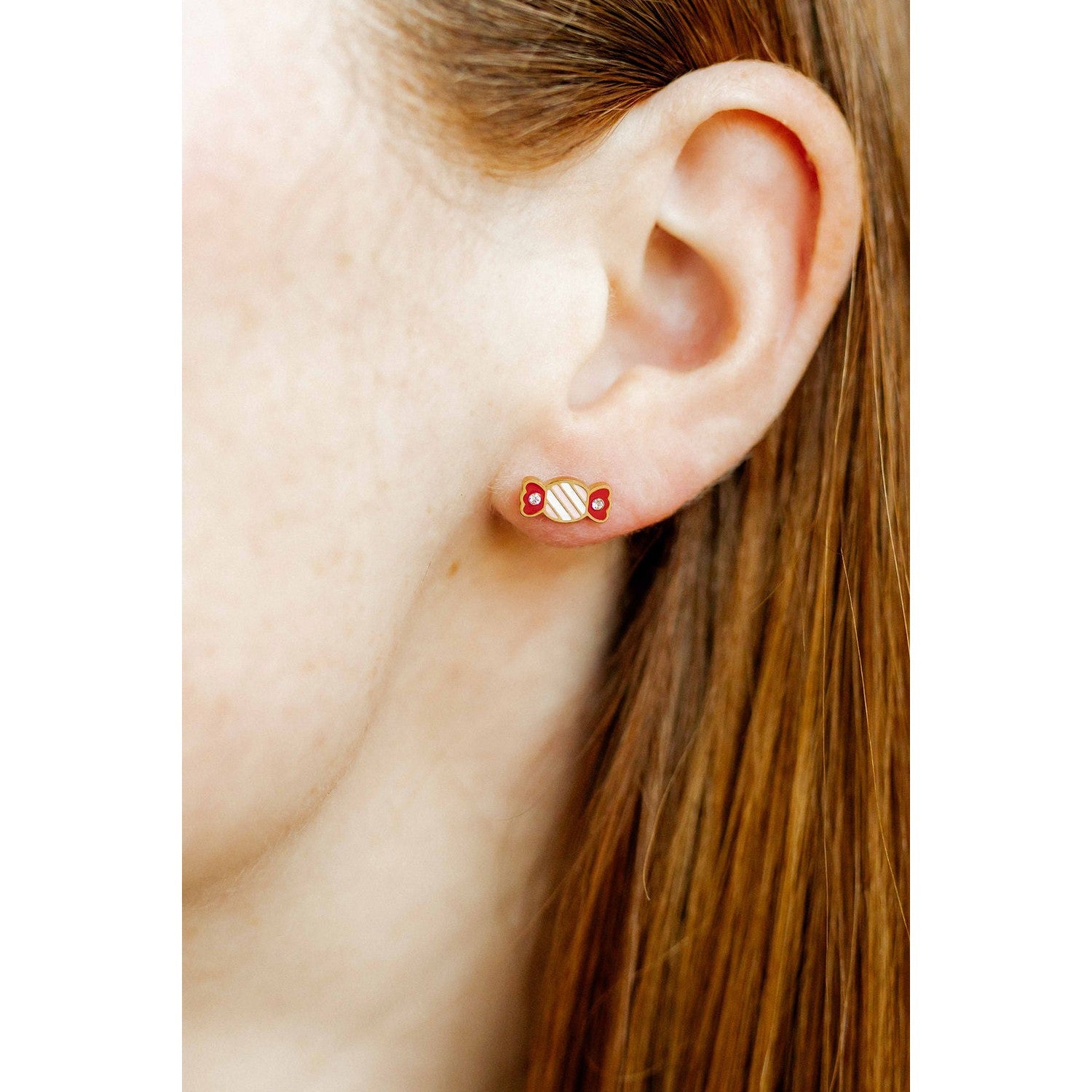 Close-up photograph of a woman's ear adorned with a gold-toned candy-shaped stud earring featuring red enamel ends, white and red diagonal stripes, and two small clear crystals