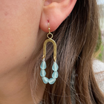 A close-up of an ear adorned with a U-shaped hammered brass earring featuring dangling seafoam green frosted glass tear-drop beads framed by brown hair