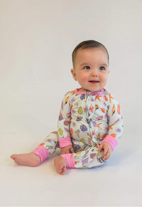 Digital photograph of a smiling infant sitting in a white studio wearing a long-sleeved romper patterned with colorful orange starfish and purple seashells featuring bright pink cuffs and a ruffled front zipper