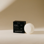 A minimalist product shot featuring a black Preston Fortifying Tea Tree Amber shampoo bar box with white text positioned next to a round white shampoo bar on a light beige surface with a subtle shadow