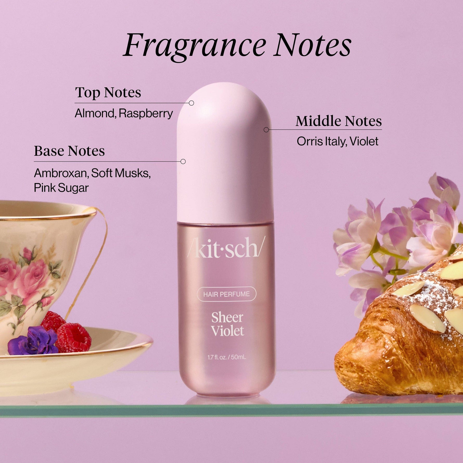 Product photograph of Kitsch Sheer Violet Hair Perfume bottle on a glass shelf, surrounded by a vintage floral teacup with raspberries, an almond croissant, and text detailing fragrance notes including Almond, Raspberry, Orris Italy, Violet, Ambroxan, Soft Musks, and Pink Sugar, all on a light purple background