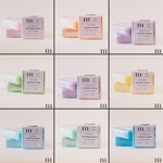 Commercial product photography for m BATH + BODY showcasing a 3x3 grid of colorful square shower bombs in pastel shades of lavender, orange, and blue with minimalist white labels and a prominent black m logo on a white background