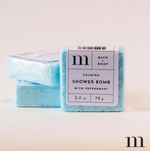 Digital photograph of m brand Calming Shower Bombs with Peppermint featuring two light blue cubes, individually wrapped, with white labels displaying the product name and weight