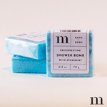 Product photograph of three bright aqua blue square M BATH + BODY Decongesting Shower Bombs with Spearmint, one in focus clearly displaying its white label with '2.5 oz 70 g' text and two others stacked behind
