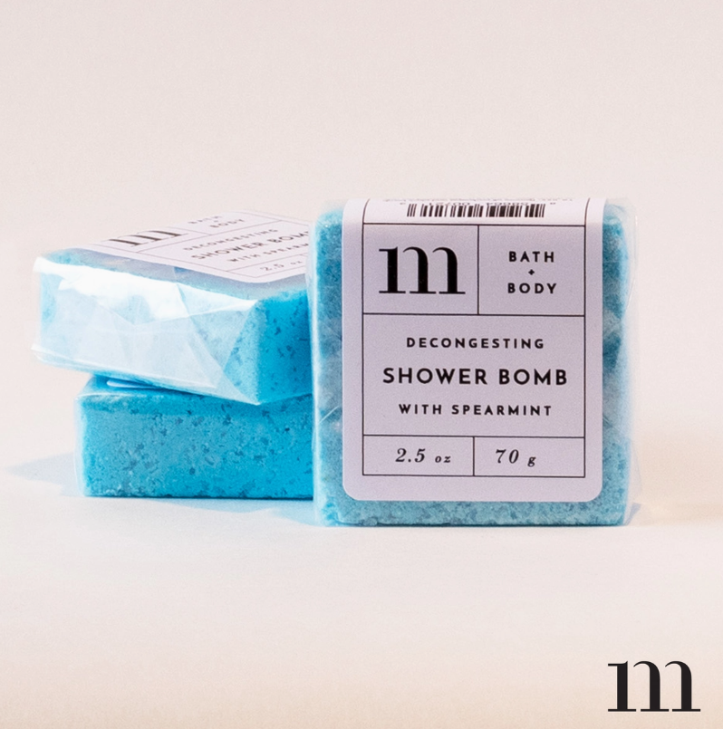 Product photograph of three bright aqua blue square M BATH + BODY Decongesting Shower Bombs with Spearmint, one in focus clearly displaying its white label with '2.5 oz 70 g' text and two others stacked behind