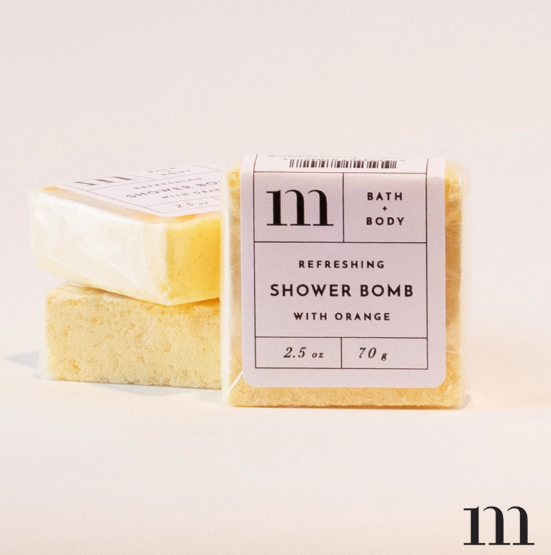 Product photograph of three square yellow "m Bath + Body Refreshing Shower Bomb with Orange" products, the front one clearly displaying its white label, with two more stacked behind it on a minimalist light background