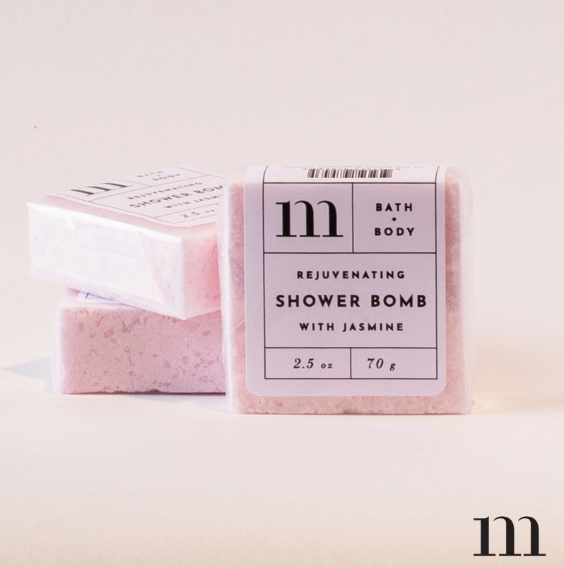Digital photograph of m BATH + BODY rejuvenating jasmine shower bombs featuring a stack of three textured pink square blocks with minimalist white labels and a bold black m logo