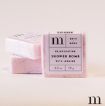 Photograph of three "m" brand Rejuvenating Shower Bombs with Jasmine, pale pink and wrapped in clear plastic with minimalist white labels featuring black text, arranged in a clean studio shot on a light background