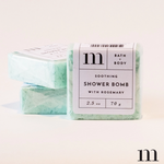 Product photography of three rectangular light green "m" brand Soothing Shower Bombs with Rosemary, the front one prominently displaying its white label detailing brand and product information in a clean studio setting
