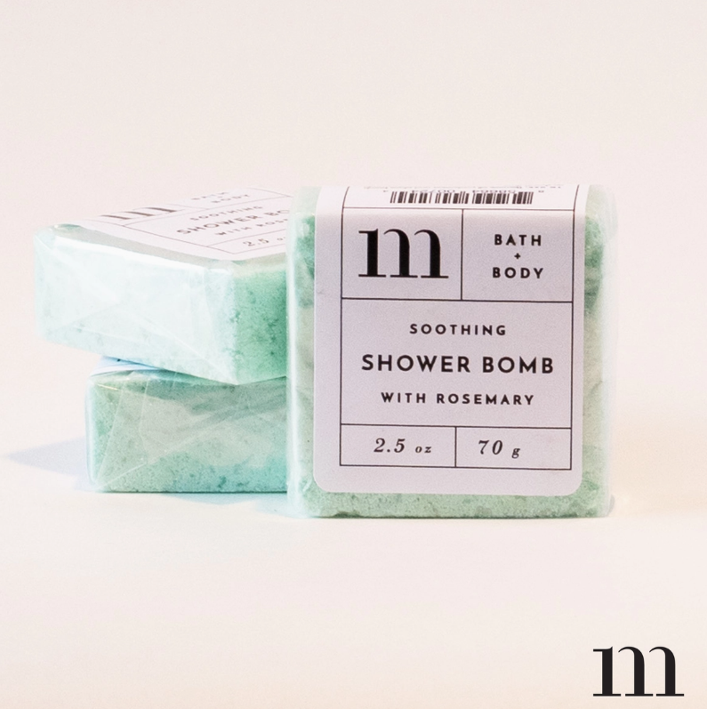 Product photography of three rectangular light green "m" brand Soothing Shower Bombs with Rosemary, the front one prominently displaying its white label detailing brand and product information in a clean studio setting