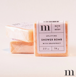 M Bath + Body product photography of three peachy-orange uplifting grapefruit shower bombs with white labels and black "m" logo on a minimal white background