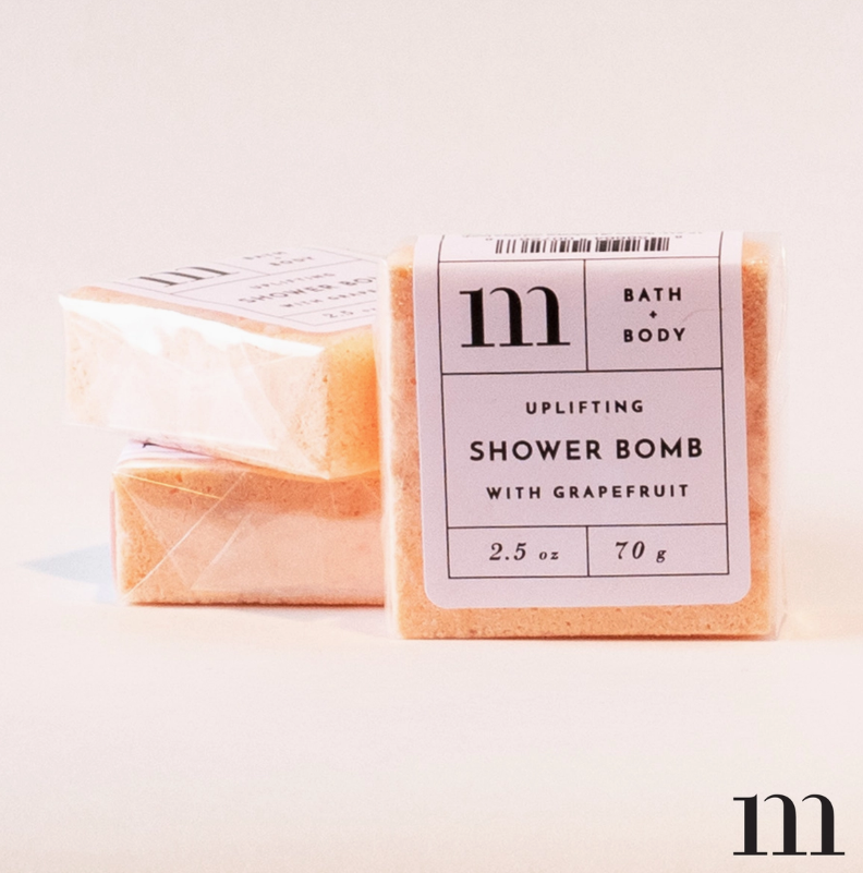 M Bath + Body product photography of three peachy-orange uplifting grapefruit shower bombs with white labels and black "m" logo on a minimal white background