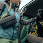 A woman with a blonde braid wearing a teal Orvis jacket and black Yeti sling bag stands at the open back of a vehicle, petting a black labrador sporting a yellow patterned bandana and green waste bag dispenser, with visible raindrops