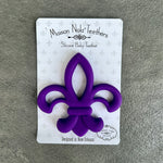 Maison Nola Teethers silicone baby teether in vibrant purple shaped as a fleur-de-lis with the brand name embossed, on white packaging featuring grey fleur-de-lis patterns, designed in New Orleans