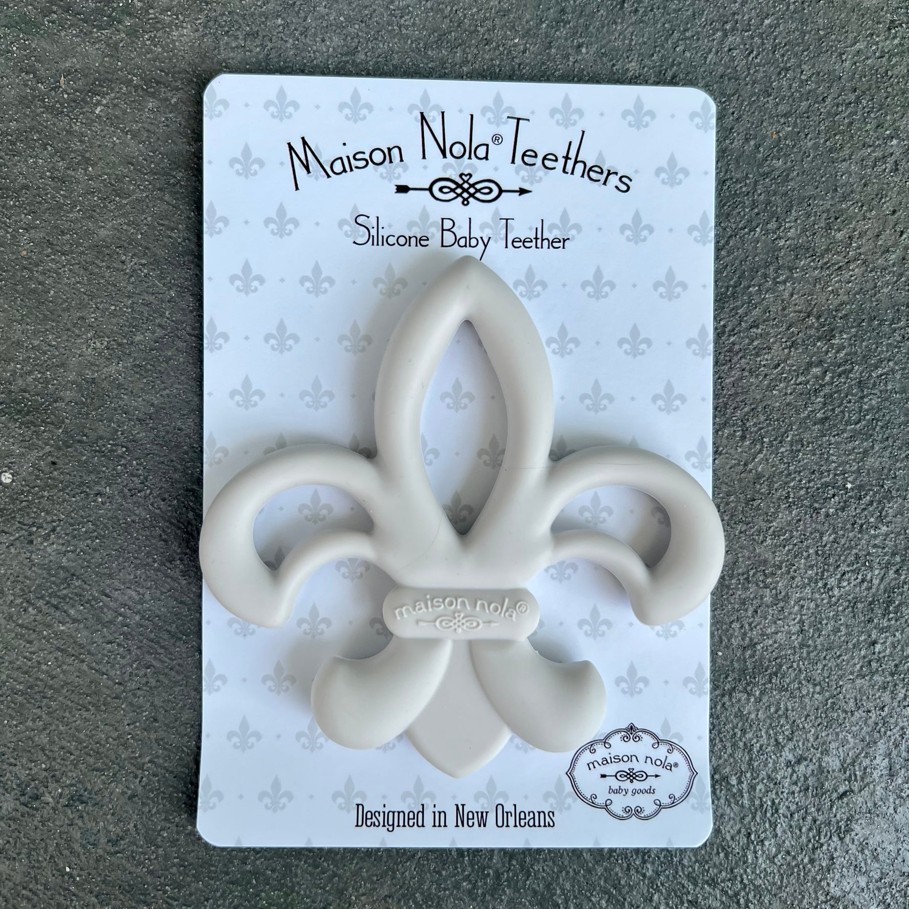 Maison Nola silicone baby teether designed in New Orleans, featuring a light gray fleur-de-lis shape on white packaging with a repeating gray fleur-de-lis pattern over a dark textured background