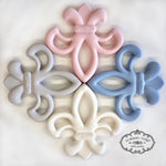 Photograph by Maison Nola Baby Goods featuring four silicone Fleur-de-lis baby teethers representing the New Orleans symbol arranged in a circular pattern in pastel pink, blue, gray, and white on a clean white background