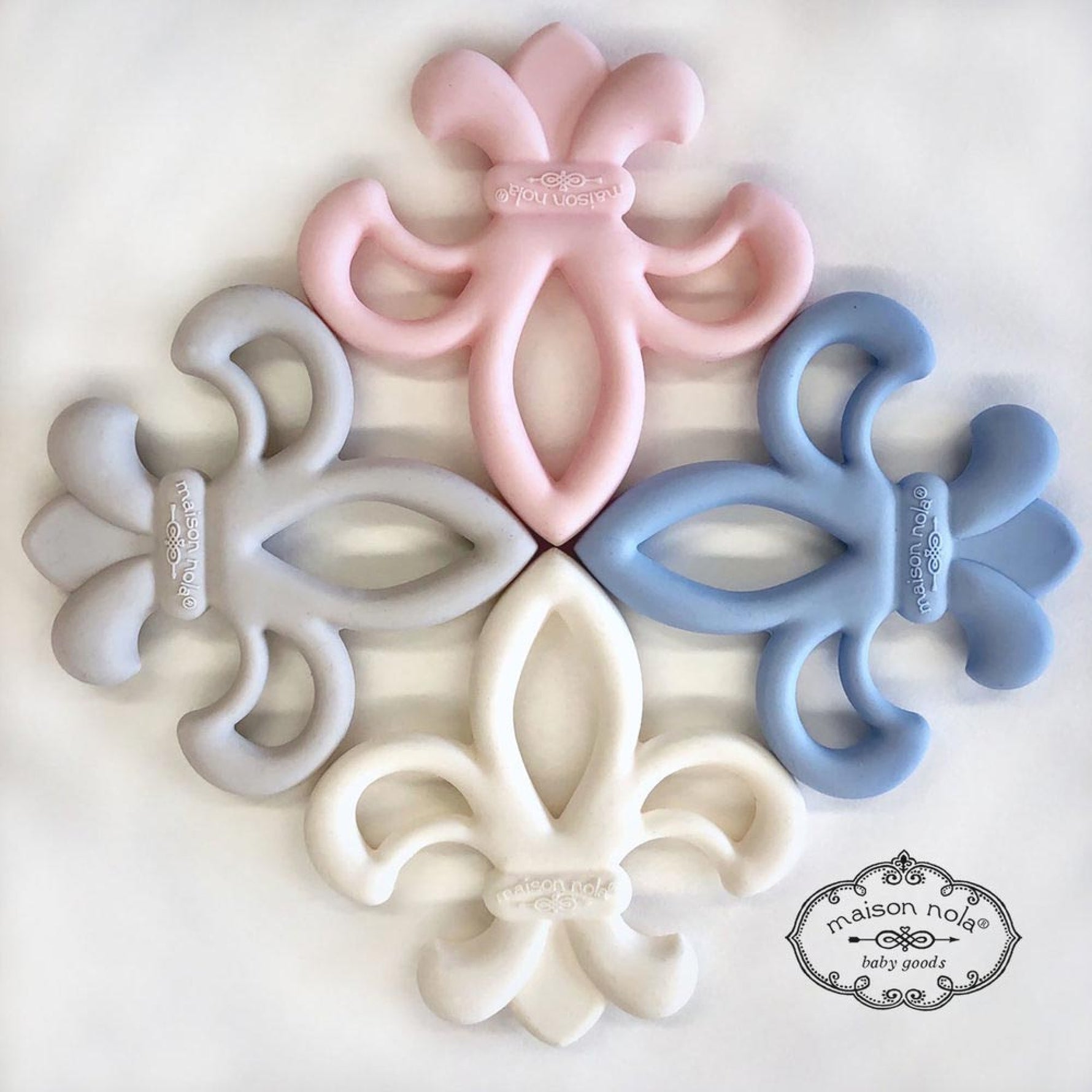 Photograph by Maison Nola Baby Goods featuring four silicone Fleur-de-lis baby teethers representing the New Orleans symbol arranged in a circular pattern in pastel pink, blue, gray, and white on a clean white background