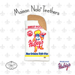 Maison Nola Teethers silicone baby product depicting an iconic Hubig's Sweet Potato Pie wrapper teether featuring its yellow baker mascot and 'New Orleans Style Pies' text, with a brown 'bite' visible against a subtle fleur-de-lis background
