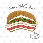 Maison Nola Teethers product is a silicone teether shaped like a New Orleans muffuletta sandwich featuring a sesame seed bun, layered meats and cheeses, and vibrant green olive salad with red pimentos, all against a subtle grey fleur-de-lis background