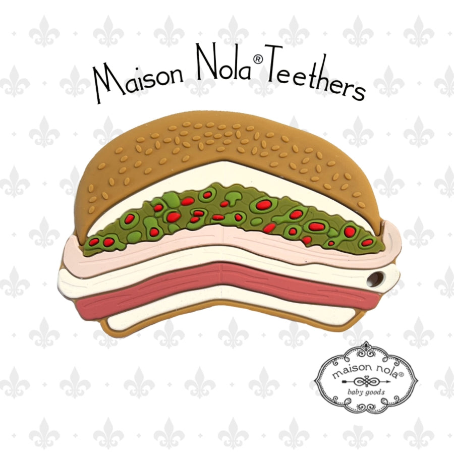 Maison Nola Teethers product is a silicone teether shaped like a New Orleans muffuletta sandwich featuring a sesame seed bun, layered meats and cheeses, and vibrant green olive salad with red pimentos, all against a subtle grey fleur-de-lis background