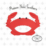 Maison Nola Teethers digital product image featuring a vibrant red silicone crab teether with distinct black eyes against a white background adorned with a subtle grey fleur-de-lis pattern