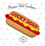 Maison Nola Teethers depicts a silicone hot dog baby teether inspired by New Orleans Lucky Dogs, featuring bold red LUCKY DOGS text, yellow mustard, and a subtle grey fleur-de-lis pattern background