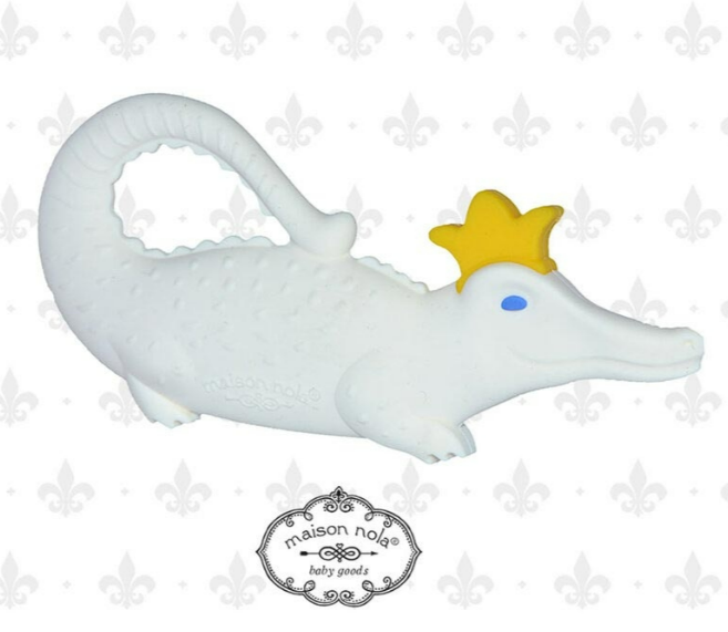Maison Nola Baby Goods silicone teether of a white alligator with bright blue eyes and a yellow crown, against a subtle fleur-de-lis pattern background suggesting New Orleans culture