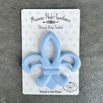 Maison Nola Teethers light blue silicone baby teether in an iconic Fleur-de-lis shape, featuring an embossed Maison Nola logo and packaged on white card with a subtle grey Fleur-de-lis pattern, designed in New Orleans