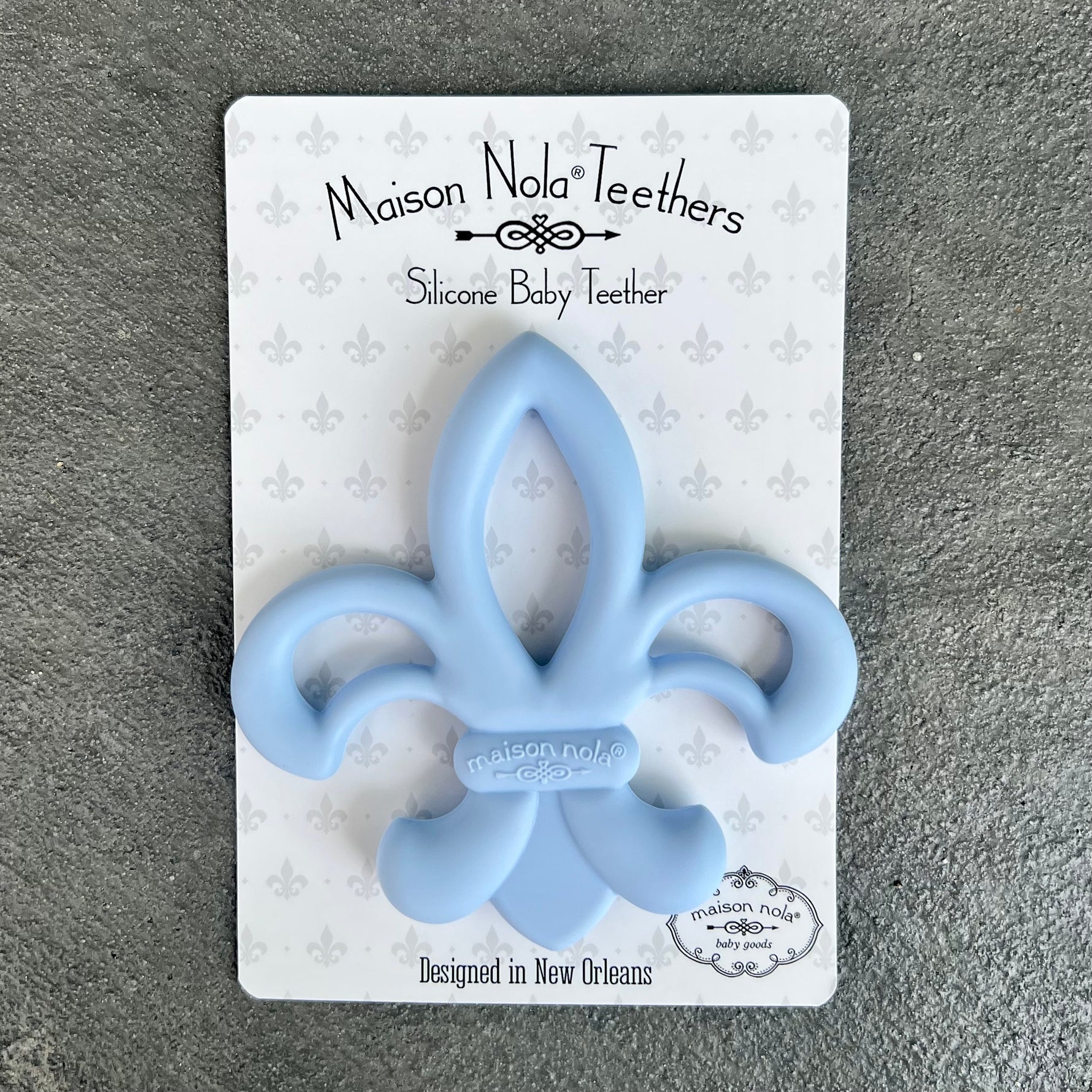 Maison Nola Teethers light blue silicone baby teether in an iconic Fleur-de-lis shape, featuring an embossed Maison Nola logo and packaged on white card with a subtle grey Fleur-de-lis pattern, designed in New Orleans