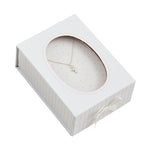 A delicate silver cross necklace displayed in a white gift box with a clear oval window, pink pinstripe details on the sides, a subtle pink polka dot interior, and a small white bow