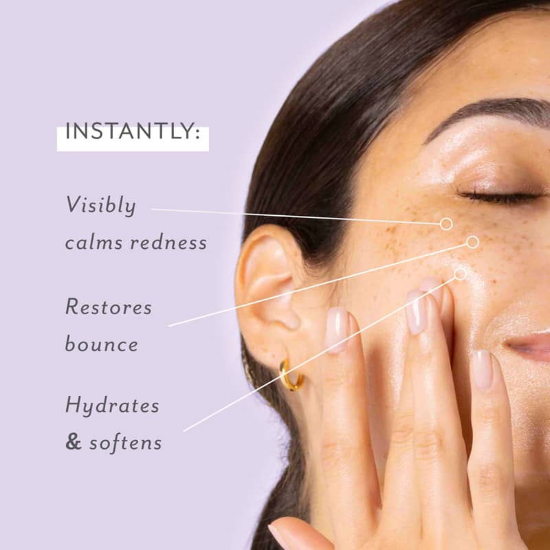 A woman with closed eyes and natural freckled skin applying skincare to her cheek, showcasing product benefits like visibly calming redness, restoring bounce, and hydrating on a lavender background with a gold hoop earring