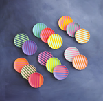 An overhead shot of a scattered assortment of small, round, vibrantly colored striped plates in various orientations on a dark gray backdrop featuring bright pink, yellow, and blue hues